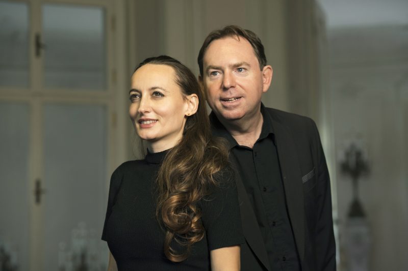 Elisabeth Schulz steht links und ist schwarz gekleidet und Emanuel Schulz steht rechts hinter ihr. Der Hintergrund ist weiß