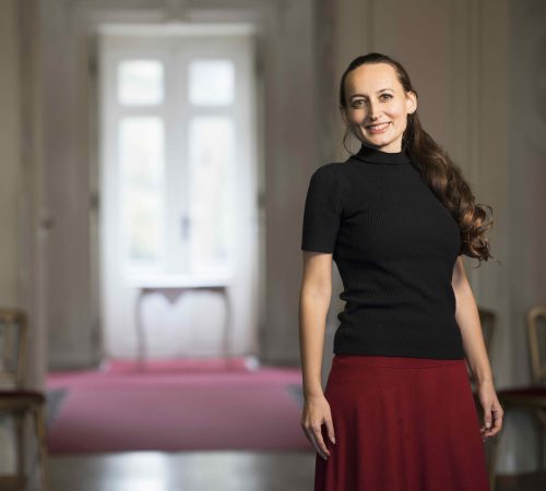 Elisabeth Schulz steht im Palais Augarten im Salon und lächelt. Sie trägt ein schwarzes Shirt und einen roten Rock. Links hinter ihr ist ein Fenster und ein roter Teppich zu sehen. Sie unterrichtet indische Musiktherapie