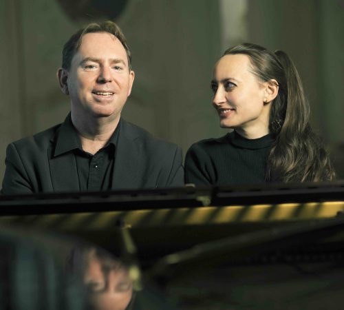 Emanuel und Elisabeth Schulz sitzen am Klavier und spielen Klavier. Emanuel sitzt links und lächelt in die Kamera. Elisabeth sitzt rechts neben ihm und lächelt ihn an, wodurch man ihr Gesicht im Profil sieht. Sie sind schwarz gekleidet. Das Klavier ist auch schwarz. Der Hintergrund ist grau. Sie spielen heilsame Musik.