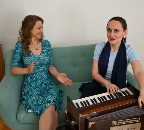 Indische Musiktherapie mit Elisabeth Schulz und einer Klientin. Die 2 Frauen sitzen am türkisen Sofa. Elisabeth spielt indisches Harmonium und singt.