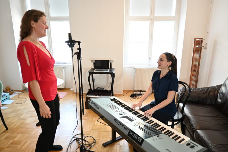 Gesangsunterricht von Elisabeth Schulz mit einer erwachsenen Schülerin. Elisabeth spielt Synthesizer und sitzt am Sessel. Ihr gegenüber steht eine blonde große Schülerin und singt in ein Mikrophon.