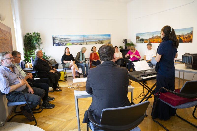 Heilsame Musik Seminar am Unum Institut mit Elisabeth Schulz und Emanuel Schulz.. 10 Leute sitzen im Halbkreis. Elisabeth Schulz steht vor den Leuten mit einem Keyboard und erklärt etwas. Emanuel sitzt neben ihr. Emanuel und Elisabeth siht man von hinten.