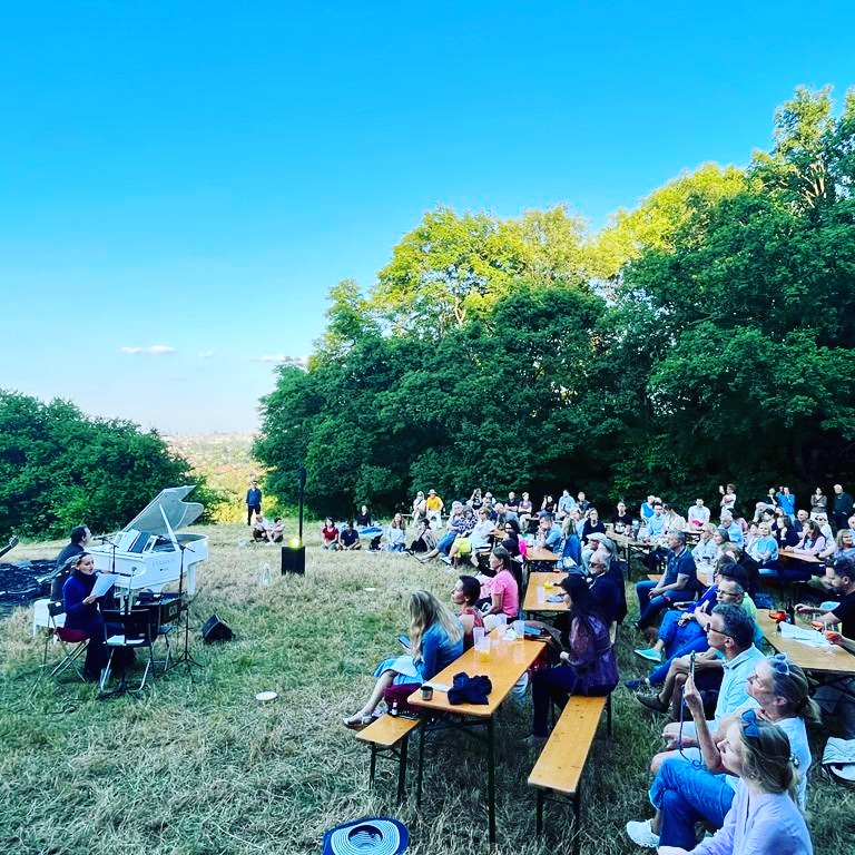 Eliem Konzert in Wien, am roten Berg. Elisabeth sitzt am Sessel mit einem Mikrophon und singt . Emanuel sitzt am Sessel und spielt Klavier auf einem weißen Flügel. Sie singen und spielen im Freien auf einer Wiese. Daneben sind Bäume und viele Zuschauer sitzen den Beiden gegenüber und hören zu.