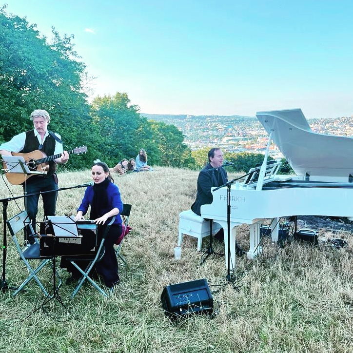 Eliem Konzert in Wien am roten Berg. Links steht Dominik Nostitz und spielt Gitarre. Daneben sitzt Elisabeth Schulz und spielt Harmonium und singt ins Mikrofon. Rechts neben ihr sitzt Emanuel Schulz am weißen Flügel, spielt Klavier und singt ins Mikrophon. Sie spielen und singen im Freien auf einer Wiese am roten Berg. Im Hintergrund sieht man die Aussicht über Wien und einen blauen Himmel.