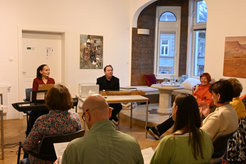 Heilsame Musik Gruppenseminar Feelharmonia mit Emanuel Schulz und Elisabeth Schulz. Mehrere Leute sitzen und hören zu. Emanuel und Elisabeth stehen vorne und spielen Musik auf indischen Instrumenten